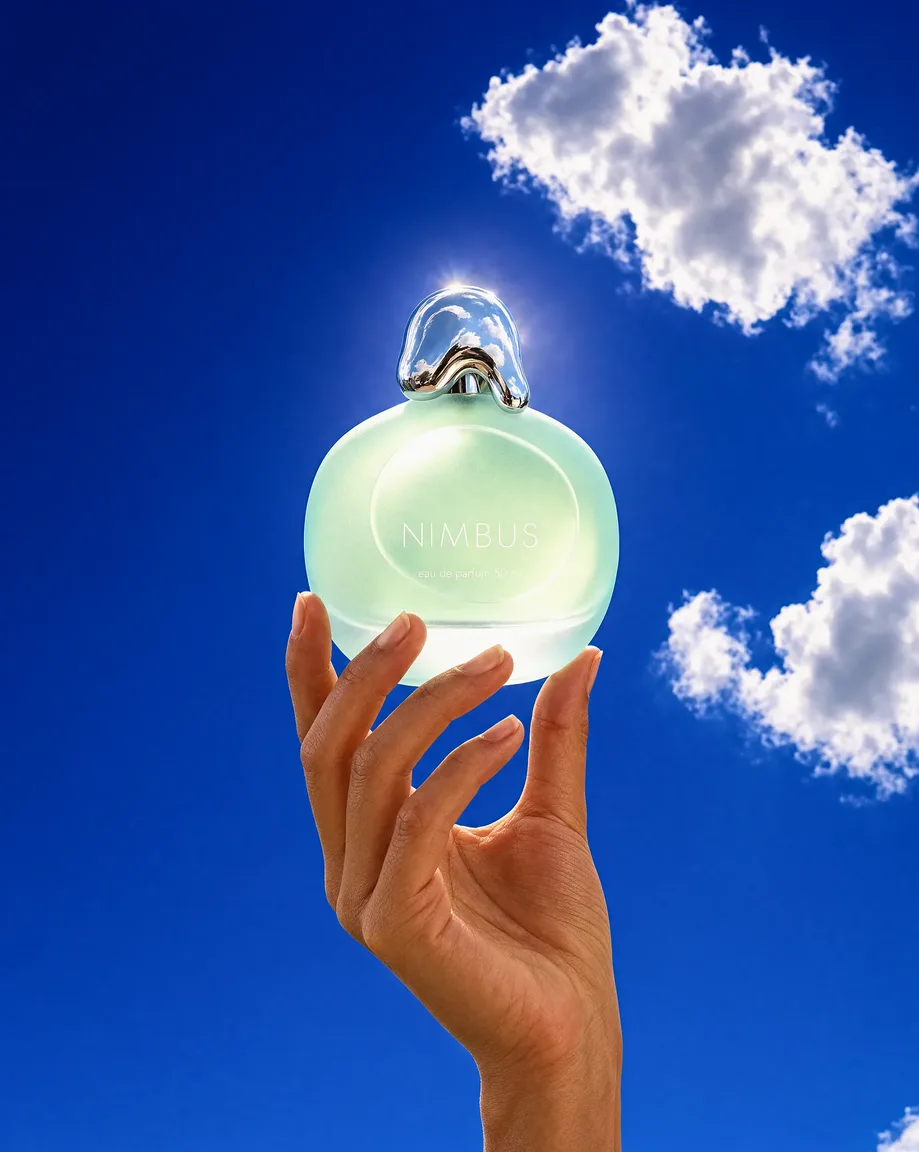 A hand raising the same perfume bottle against a cobalt summer sky with white clouds